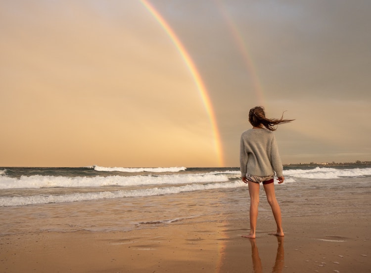 young woman standing on beach looking at two rainbows