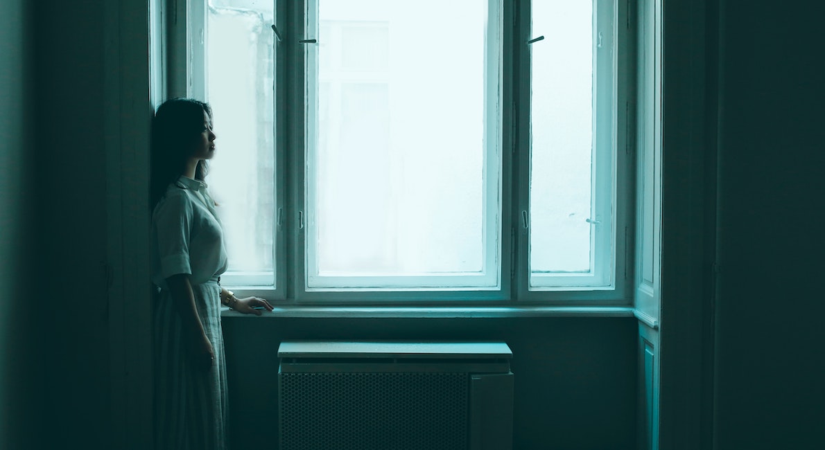 young woman looking out a window in a darkened room.